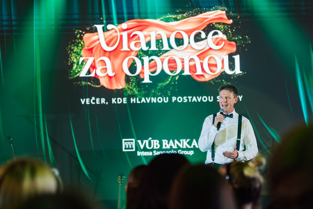 Vystupujúci s mikrofónom pred obrazovkou s nápisom Vianoce za oponou