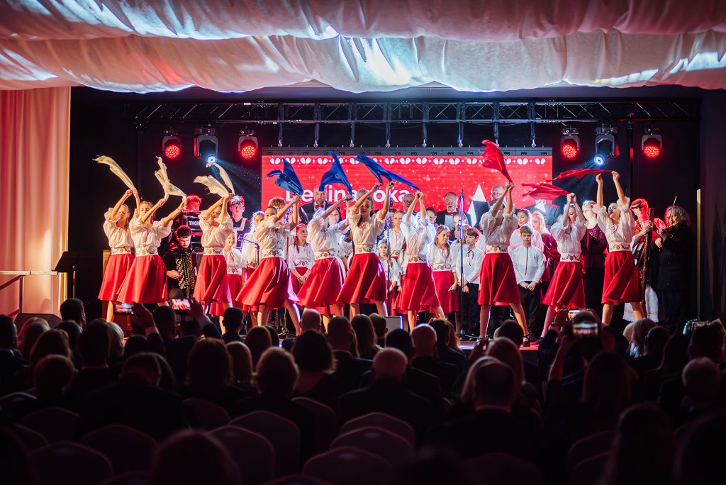 Folklórna skupina tancuje na pódiu pred plnou sálou, v rukách červené šatky