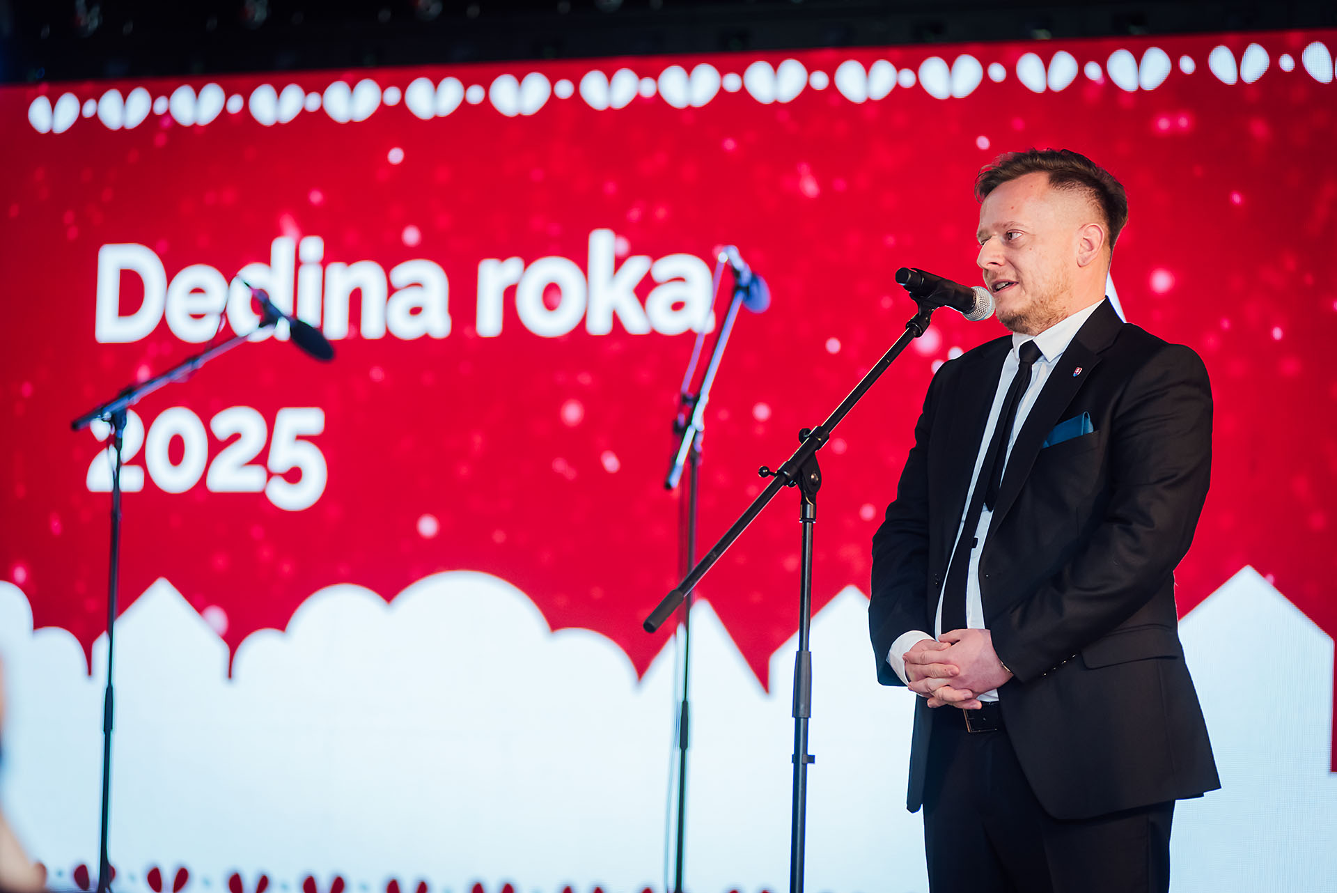 Rečn&iacute;k pri mikrof&oacute;ne pred obrazovkou s n&aacute;pisom Dedina roka 2025